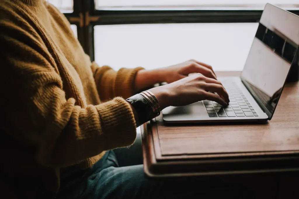 Person in a yellow sweater typing on a laptop at a desk near a window.
